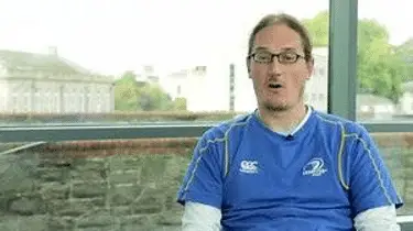 A man wearing glasses and a blue shirt sits on a bench, looking thoughtfully into the distance.
