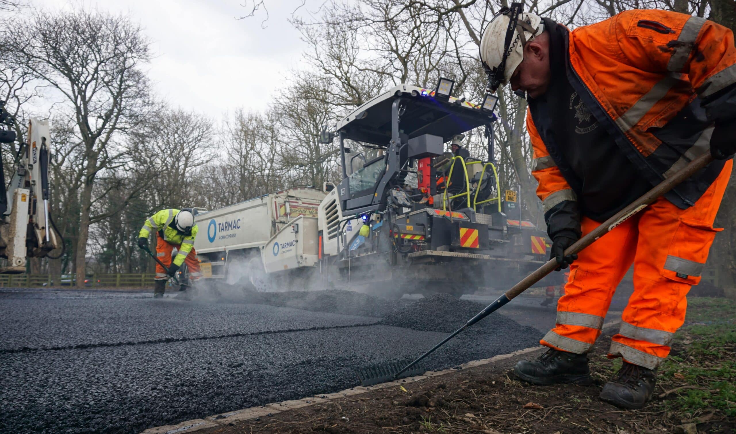 A worker applies asphalt to a road surface, wearing safety gear and using a tool for smooth application.